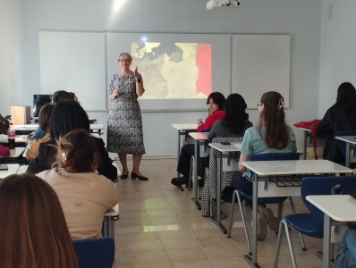 The photo shows the room where classes are held. The teacher explains something to a group of students. In the background there is a slide with a presentation.. Click to zoom the picture.