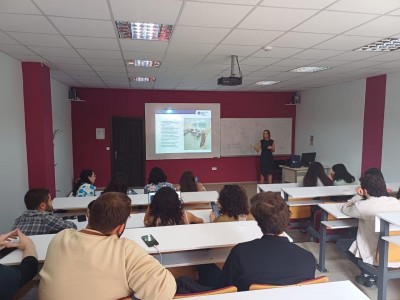 The photo shows the room where classes are held. The teacher explains something to a group of students. In the background there is a slide with a presentation. Click to zoom the picture.