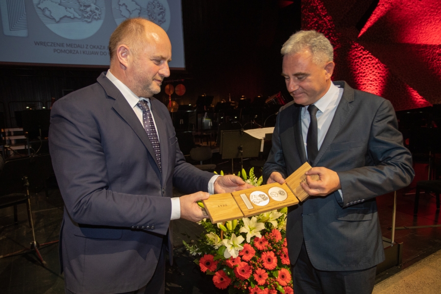 Profesor Jarosław Kłaczkow (z prawej) odbiera z rąk Marszałka Województwa Kujawsko-Pomorskiego pamiątkowy medal upamiętniający powrót Pomorza i Kujaw do wolnej Polski w 1920 roku fot.Urząd Marszałkowski Województwa Kujawsko-Pomorskiego Na scenie sali koncertowej na toruńskich Jordankach stojący po prawej stronie profesor Jarosław Kłaczkow odbiera z rąk stojącego po lewej stronie Marszałka Województwa Piotra Całbeckiego pamiątkowy medal (stylizowane na liczbę sto metalowe odlewy umieszczone w trójdzielnej drewnianej podstawie). Obaj panowie ubrani na galowo, w tle kosz z bukietem kwiatów, puste krzesła i pulpity dla orkiestry.