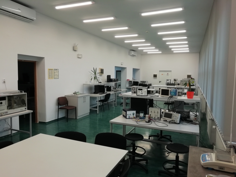 Laboratorium główne fot.dr hab. D. Piątkowski, prof. UMK Fotografia przedstawia duże laboratorium. Ściany pomalowane na biało, posadzka zielona. Po lewej stronie widoczne otwarte drzwi. Na pierwszy planie widoczny fragment szarego stołu. Za nim szare biurka laboratoryjne z nadstawkami ustawione w rzędzie, przy nich stoją krzesła i taborety. Na biurkach stoi specjalistyczna aparatura badawcza, m.in. oscyloskopy, zasilacze, multimetry i komputery. Po prawej stronie rząd okien zasłoniętych szarymi żaluzjami. Widoczny jest biały sufit, na tle którego wyróżniają się dwa rzędy jasnych lamp. Na ścianie po lewej stronie widoczne dwa klimatyzatory, instrukcje BHP, oświetlenie awaryjne oraz punkt dostępu do sieci Eduroam.