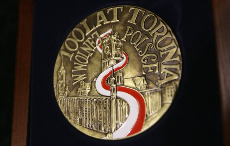 Medal 100-lecia Torunia w Wolnej Polsce fot.Adam Zakrzewski (UM Toruń) Zdjęcie okrągłego medalu, najpewniej z brązu, z wizerunkem toruńskiego rausza staromiejskiego, z biało-czerwoną flagą spływającą serpentynami z masztu na wieży zegarowej do pomnika Mikołaja Kopernika i napisem: 100 LAT TORUNIA W WOLNEJ POLSCE