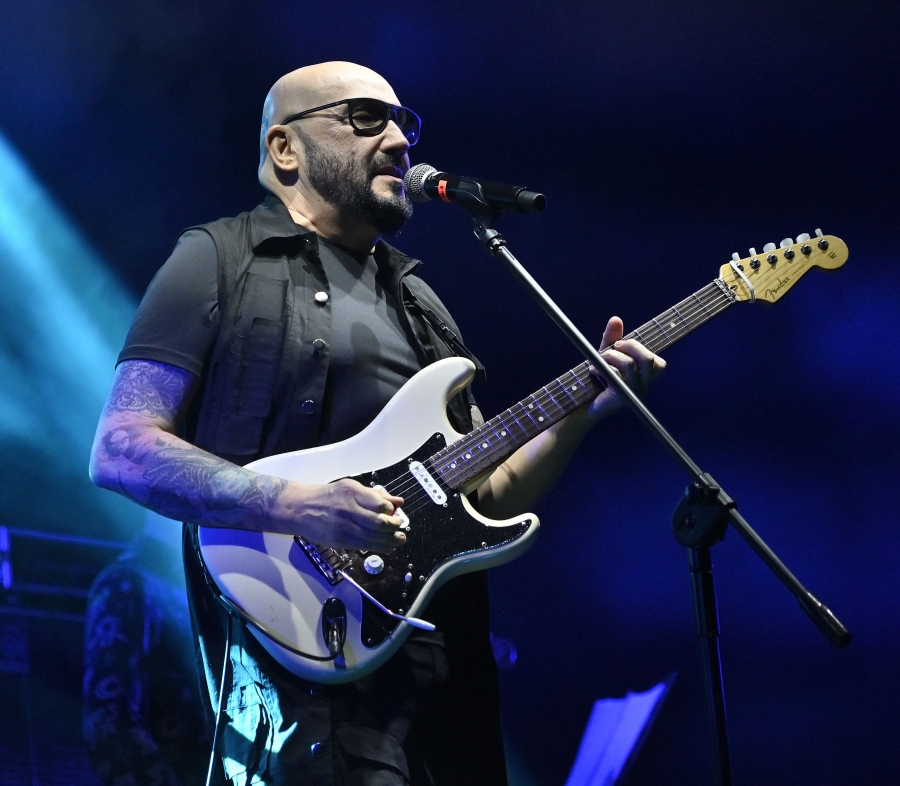 fot.Tomasz Dorawa Bohater rozmowy stojący na scenie. Ma założone ciemne okulary, ciemny t-shirt i koszulę z krótkim rękawe. Widoczny tatuaż na prawym przedramieniu. Muzyk gra na gitarze i śpiewa do mikrofonu umieszczonego na statywie. Ciemne tło ze smugą światła scenicznego za plecami postaci.