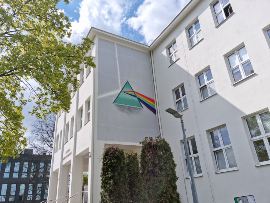 Pryzmat fot.prof. dr hab. Ireneusz Grabowski Biała ściana budynku, na której znajduje się duży mural przedstawiający pryzmat rozszepiający światło białe na barwy podstawowe.