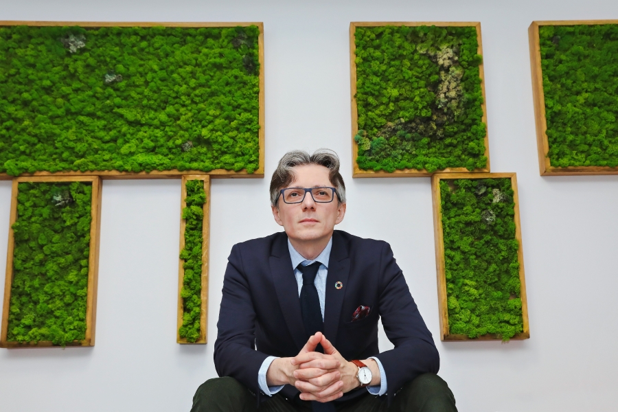fot.Andrzej Romański Portret bohatera wywiadu patrzącego przed siebie, siedzącego naprzeciw nas; jest ubrany elegancko (marynarka, koszula, krawat, zegarek); tuż za nim biała ściana z ujętymi w drewniane ramy zielonymi kompozycjami mchu.