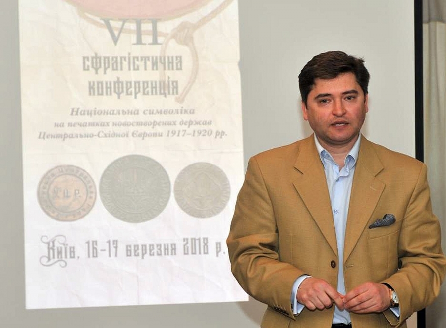 dr Vitaliy Perkun fot.sigillum.com.ua Młody mężczyzna w beżowej marynarce stojący na tle ekranu projekcyjnego