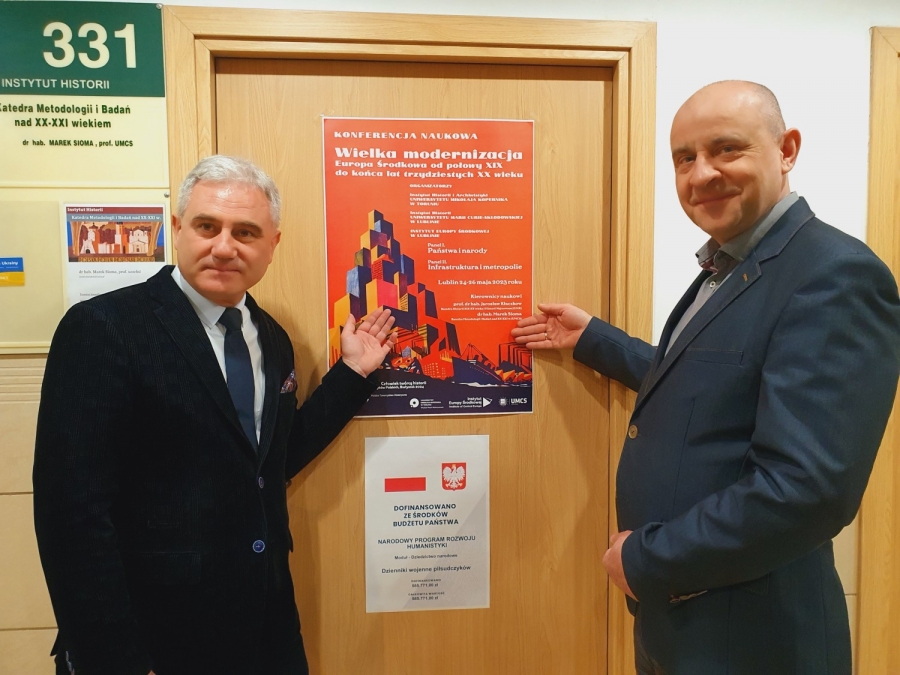 Kierownicy konferencji "Wielka modernizacja" - od lewej: prof. Jarosław Kłaczkow, prof. Marek Sioma fot.materiały organizatorów Dwóch mężczyzn rękoma wskazuje wiszący na drzwiach plakat informujący o konferencji.