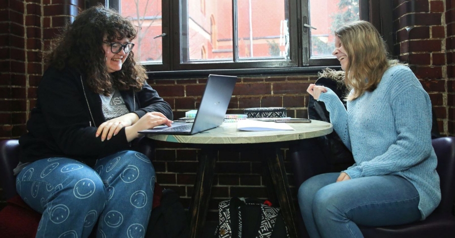 obrazek: Tasks of the tutor will include: preparing and implementing classes, setting up and operating virtual meetings and possibly consulting with those taking the course photo:Andrzej Romański Two girls sitting around the table, they are smiling, one of them has a laptop in a front of her