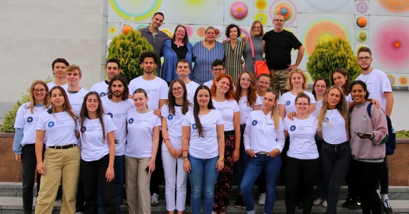 Photo description - in the foreground smiling students in two rows dressed in white t-shirts with the NCU logo. Lecturers in the third row. The photo was taken against the background of the decorative façade of the Aula UMK with the system of planets.