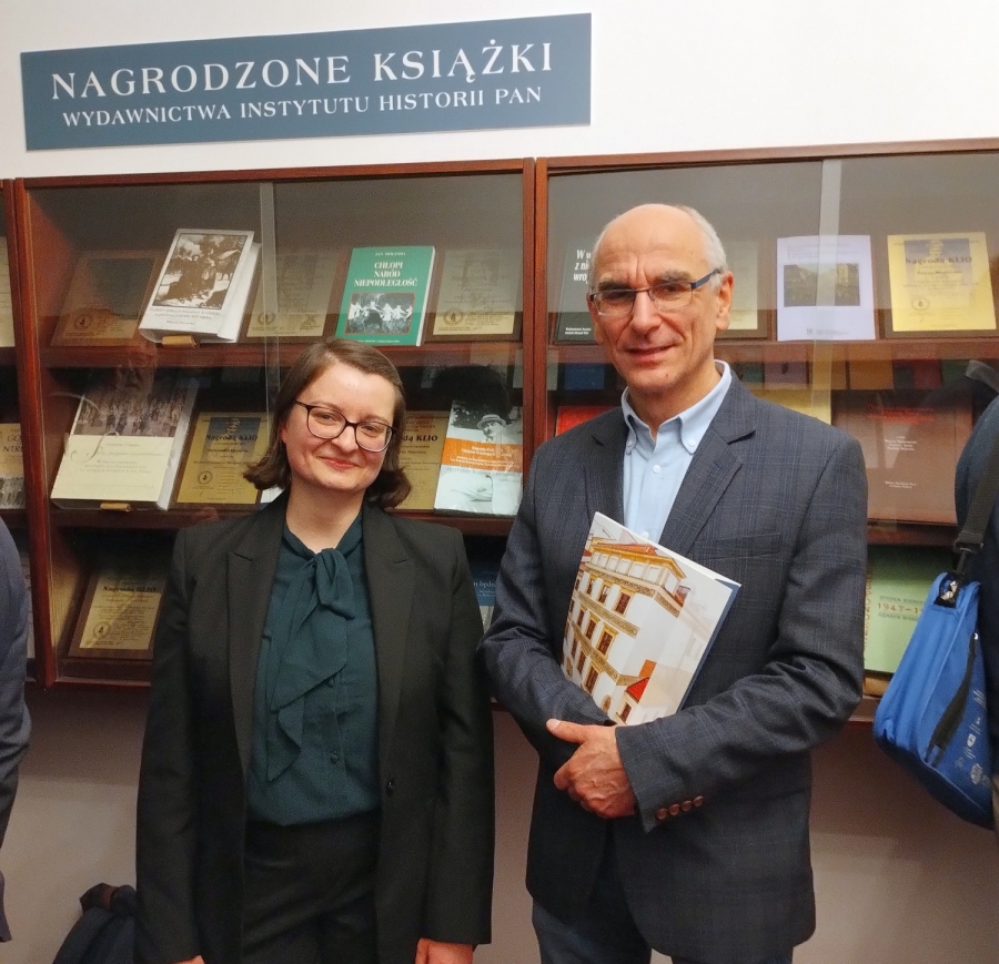 Od lewej: Hanna Rajfura, sekretarz redakcji "Studiów Źródłoznawczych" i prof. Piotr Oliński, laureat nagrody. fot.nadesłane Na tle oszklonej gabloty z nagrodzonymi książkami pozują do zdjęcia kobieta (z lewej) i mężczyzna, oboje noszą okulary.