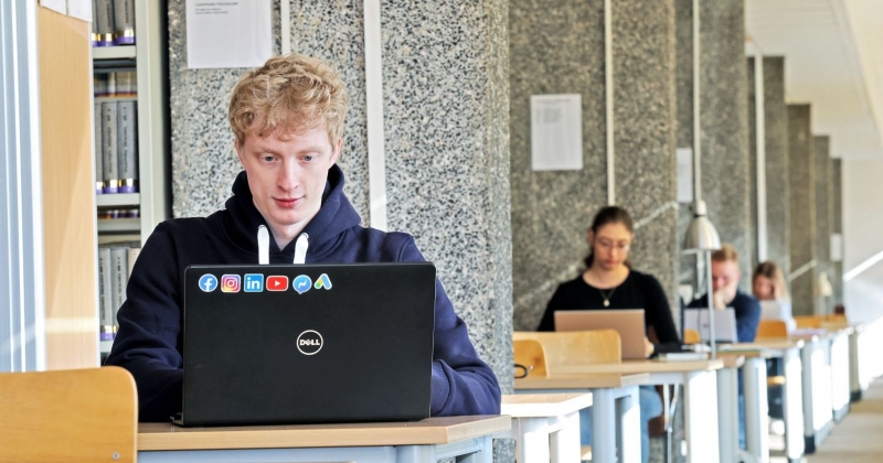 Studenci i studentki w bibliotece siedzą przy biurkach z laptopami, po lewej stronie widać również fragment regału z książkami