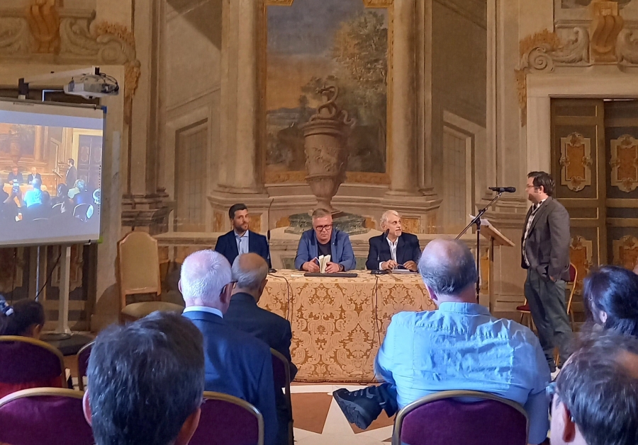 Profesor Marcin Pawlak (w środku) przy stole prezydialnym konferencji. fot.nadesłane W sali ozdobionej freskami uczestnicy konferencji: słuchacze odwróceni plecami, przed nimi stół prezydialny, przy którym siedzi trzech mężczyzn, po prawej stronie stoi prelegent przy mikrofonie.