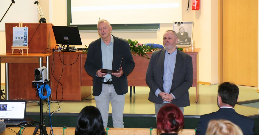 Seminatium poświęcone pamięci prof. Kazimierza Żurowskiego fot.Dorota Bienias, Ryszard Kaźmierczak, Marcin Weinkauf Na zdjęciu dwóch mężczyzn w garniturach stoi przed publicznością w sali wykładowej, jeden z nich trzyma otwartą teczkę i przemawia, w tle widoczny jest ekran, biurko z komputerem oraz czarno-biała fotografia starszego mężczyzny.