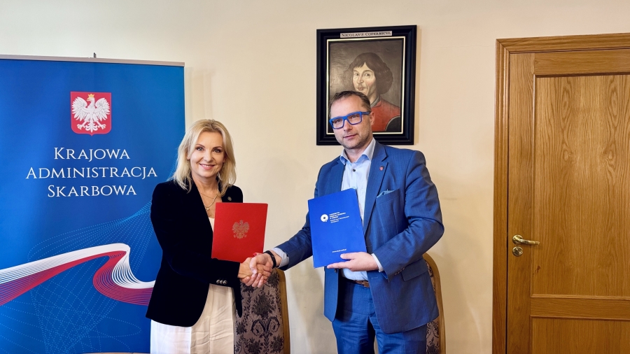 Podpisanie porozumienia odbyło się na Wydziale Nauk Ekonomicznych i Zarządzania. Dwoje ludzi, kobieta i mężczyzna, stoi w pomieszczeniu, podają sobie ręce i trzymają teczki ? kobieta czerwoną z godłem Polski, mężczyzna niebieską z logo instytucji. Za nimi widoczny jest niebieski baner z napisem ?Krajowa Administracja Skarbowa? oraz obraz przedstawiający Mikołaja Kopernika. Po prawej stronie widać drewniane drzwi.