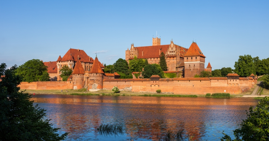 Zamek w Malborku fot.źródło: Canva amek krzyżacki w Malborku ? widok na masywną ceglaną warownię z wieżami i murami obronnymi, odbijającą się w wodach rzeki Nogat w słoneczny dzień.