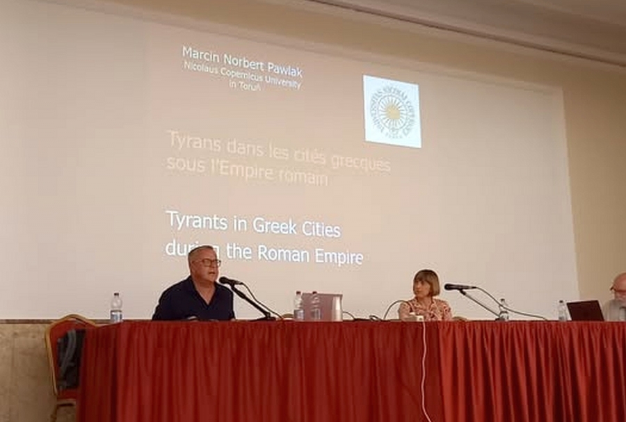 Prof. Marcin Pawlak w trakcie wygłaszania refereatu podczas Symposium Peregrinum 2025 fot.nadesłane Dwójka osób - mężczyzna i kobieta - siedzą na podwyższeniu za stołem przykrytym czerwonym suknem; w tle ekran z tytułowym slajdem prezentacji.