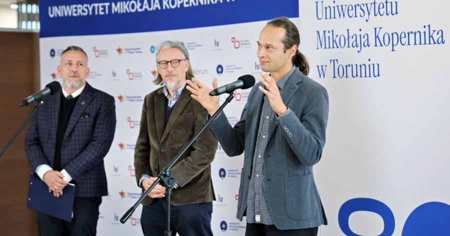 Rozpoczęciu projektu towarzyszyła konferencja prasowa w Auli UMK fot.fot. Andrzej Romański Trzech mężczyzn stoi na tle ścianki promocyjnej UMK. Przed nimi mikrofony na statywach. Jeden z mężczyzn żywiołowo gestykuluje