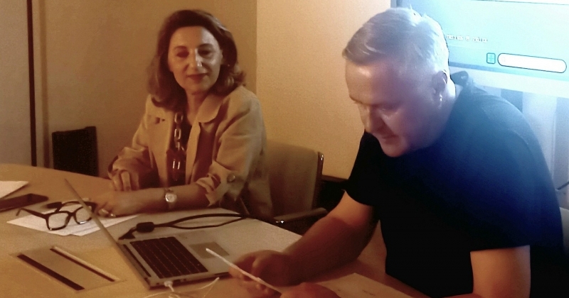 W zaciemnionej sali rozjaśnionej blaskiem padajacym z dużego ekarnu telewizyjnego, dwie ososby - kobieta i mężczyzna - siedzą przy stole, mężczyzna czyta z kartki.