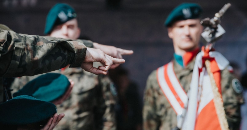 Zdjęcie z przysięgi wojskowej - widać dłonie żołnierzy wskazujące na sztandar, w tle dwóch umundurowanych żołnierzy