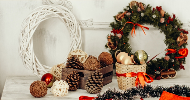 Na zdjęciu znajduje się świąteczna aranżacja z wiklinowymi i zielonymi wieńcami, szyszkami, ozdobnymi kulami i czerwonymi wstążkami ułożonymi na białym stole.