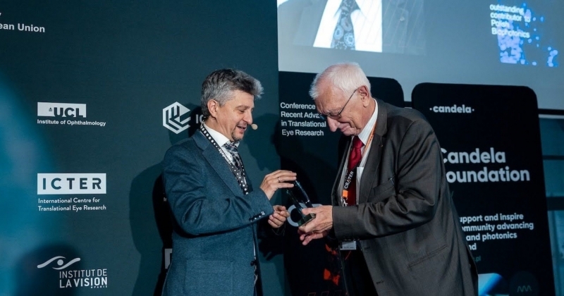 Zdjęcie przedstawia scenę z konferencji naukowej lub medycznej, na której prof. dr hab. Maciej Wojtkowski wręcza prof. dr. hab. Andrzejowi Kowalczykowi statuetkę. Prof. dr hab. M. Wojtkowski stoi przodem do prof. dr. hab. A. Kowalczyka i lekko bokiem do obiektywu. Jest to mężczyzna w średnim wieku, z krótkimi, szpakowatymi włosami. Ubrany jest w elegancki, ciemnogranatowy garnitur i krawat. Na szyi ma zawieszoną smycz identyfikacyjną. Właśnie przekazuje statuetkę do rąk drugiego mężczyzny. Uśmiecha się. Prof. dr hab. A. Kowalczyk jest starszy, ma siwe włosy i okulary. Ubrany jest w ciemny, białą koszulę i czerwony krawat. Na szyi ma również zawieszoną smycz z identyfikatorem. Pochyla się nieco do przodu, by przyjąć nagrodę. Uśmiecha się łagodnie. Obaj mężczyźni trzymają w dłoniach małą, przezroczystą statuetkę, która wygląda na wykonaną ze szkła lub akrylu. Scena rozgrywa się na tle ciemnej, profesjonalnej ściany wystawowej, pełnej logotypów i napisów związanych z konferencją. Zdjęcie ma uroczysty i pozytywny charakter, uwieczniając moment wręczenia prestiżowej nagrody podczas międzynarodowej konferencji naukowej. Obaj mężczyźni wydają się zadowoleni i honorowi.