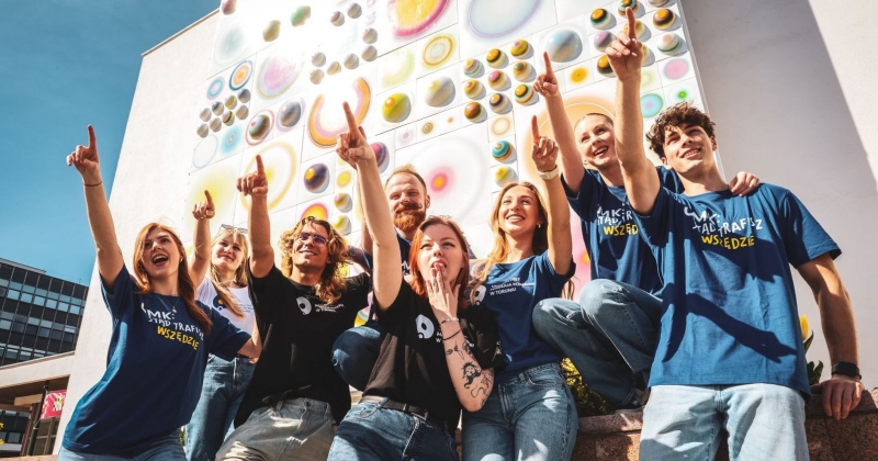 8-osobowa grupa studentek i studentów w koszulkach z napisem "Stąd traficz wszędzie" lub z logo UMK stoi na tle Auli UMK i palcem wskazuje w kierunku nieba