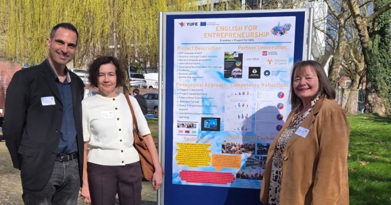 This photo was taken in a park. In the foreground is a stand with a large conference poster. Next to the poster stand three smiling people, participants of the "English for Entrepreneurship" project.