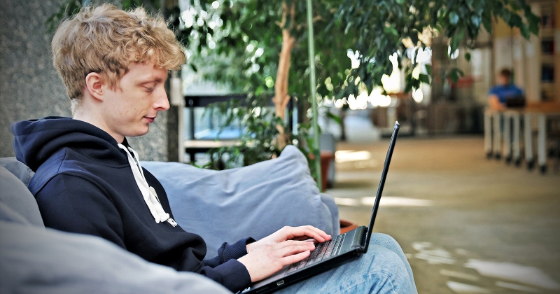 Student siedzi na kanapie z laptopem na kolanach i patrzy w ekran