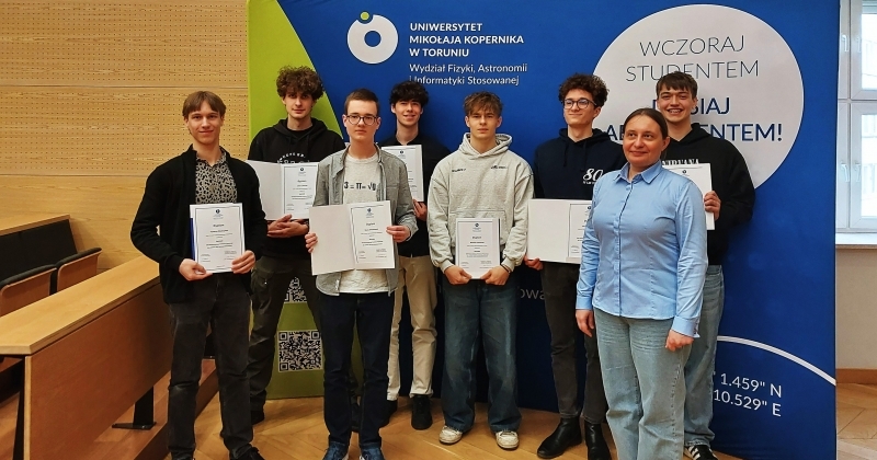 Osoby stojące i trzymające dyplomy w Audytorium Aleksandra Jabłońskiego na tle ścianki z logo WFAiIS