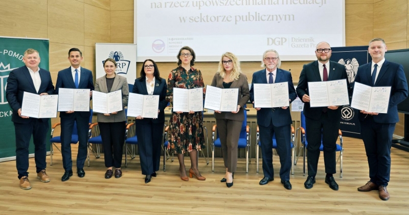Dziewięć osób, które podpisały deklarację, stoją z egzemplarzami dokumentów. Za nimi wyświetla się slajd z prezentacji, na którym napisano "Podpisanie Deklaracji Toruńskiej na rzecz upowszechnienia mediacji w sektorze publicznym"