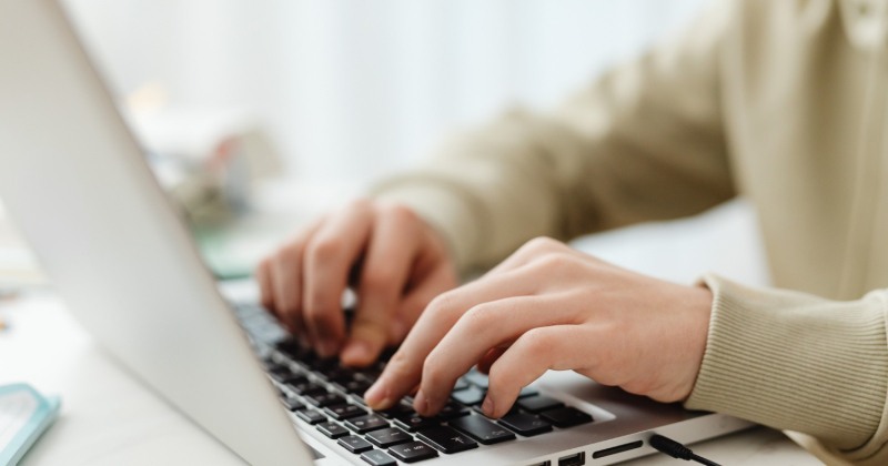Zbliżenie na dłonie osoby piszącej na klawiaturze laptopa stojącego na biurku w jasnym, dobrze oświetlonym pomieszczeniu.