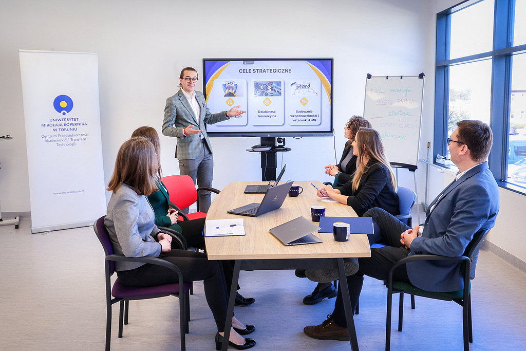 Five individuals at a table watching a presentation delivered by a person standing next to a large screen [photo: Andrzej Romański]