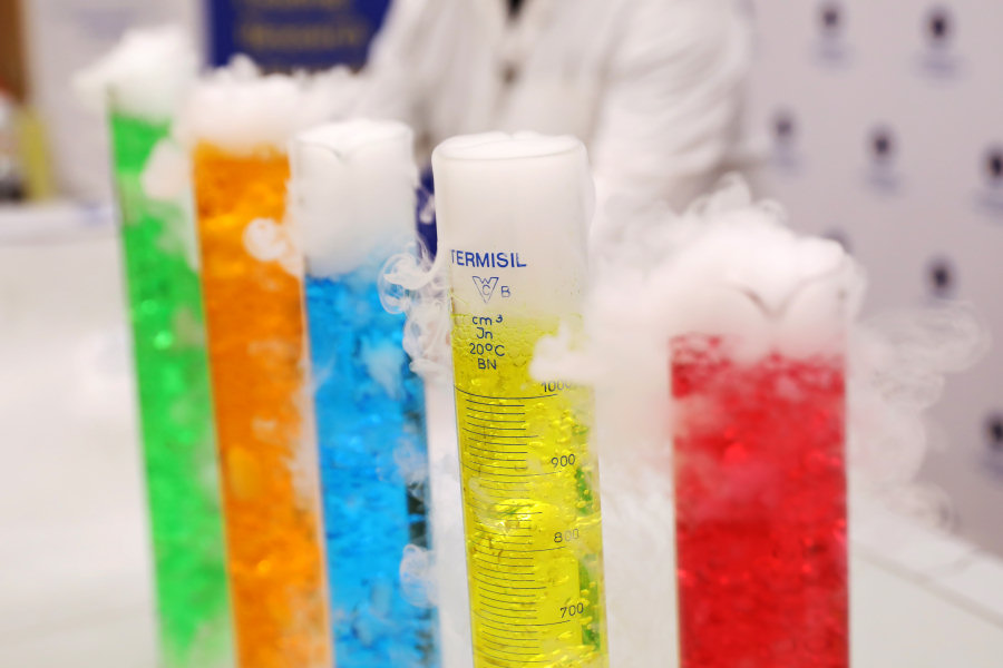 Pięć szklanych cylindrów laboratoryjnych wypełnionych kolorowymi płynami (kolejno: zielony, pomarańczowy, niebieski, żółty, czerwony). Z naczyń wydobywa się dym, co wskazuje na trwający pokaz chemiczny. W tle widoczna postać osoby w białym fartuchu oraz fragment oznakowania uczelni. [fot. Andrzej Romański]