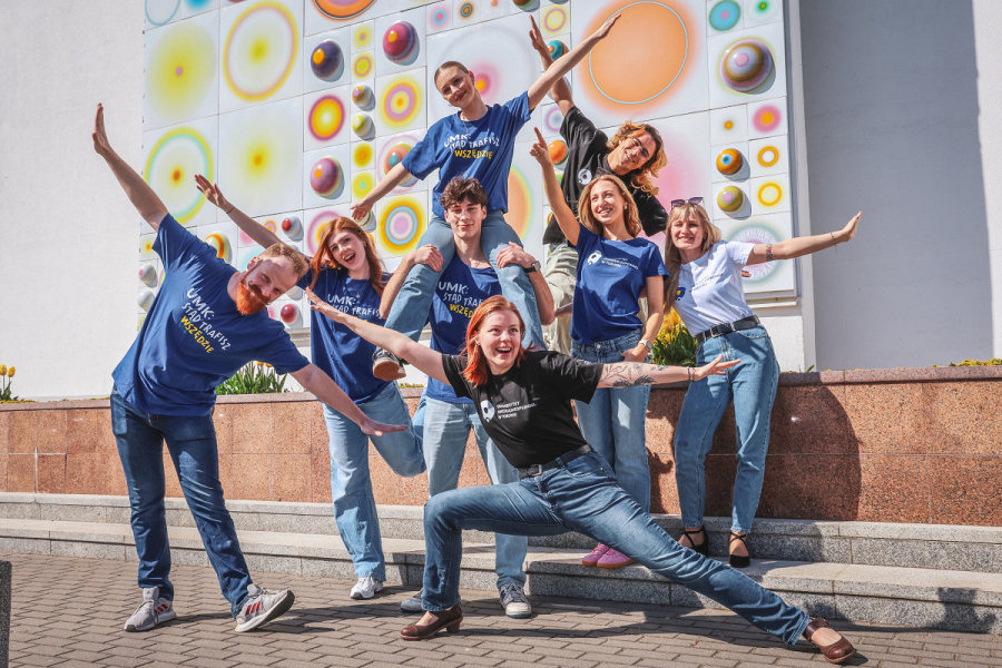 Grupa siedmiu młodych osób w koszulkach z logo UMK pozuje przed ścianą ozdobioną kolorowymi, wypukłymi elementami. Studenci wykonują dynamiczne gesty z szeroko rozłożonymi ramionami, jedna osoba znajduje się na ramionach kolegi. [fot. Andrzej Romański]