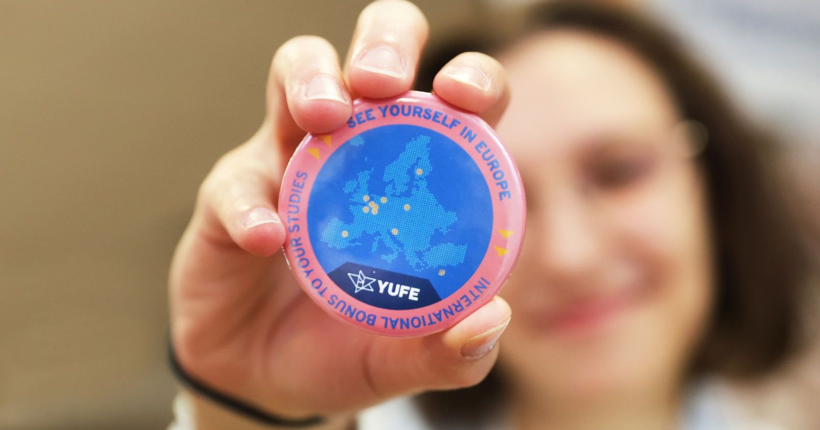 Przypinka z logo YUFE i mapą Europy trzymana w dłoni osoby, kt&oacute;rej twarz jest rozmyta na drugim planie