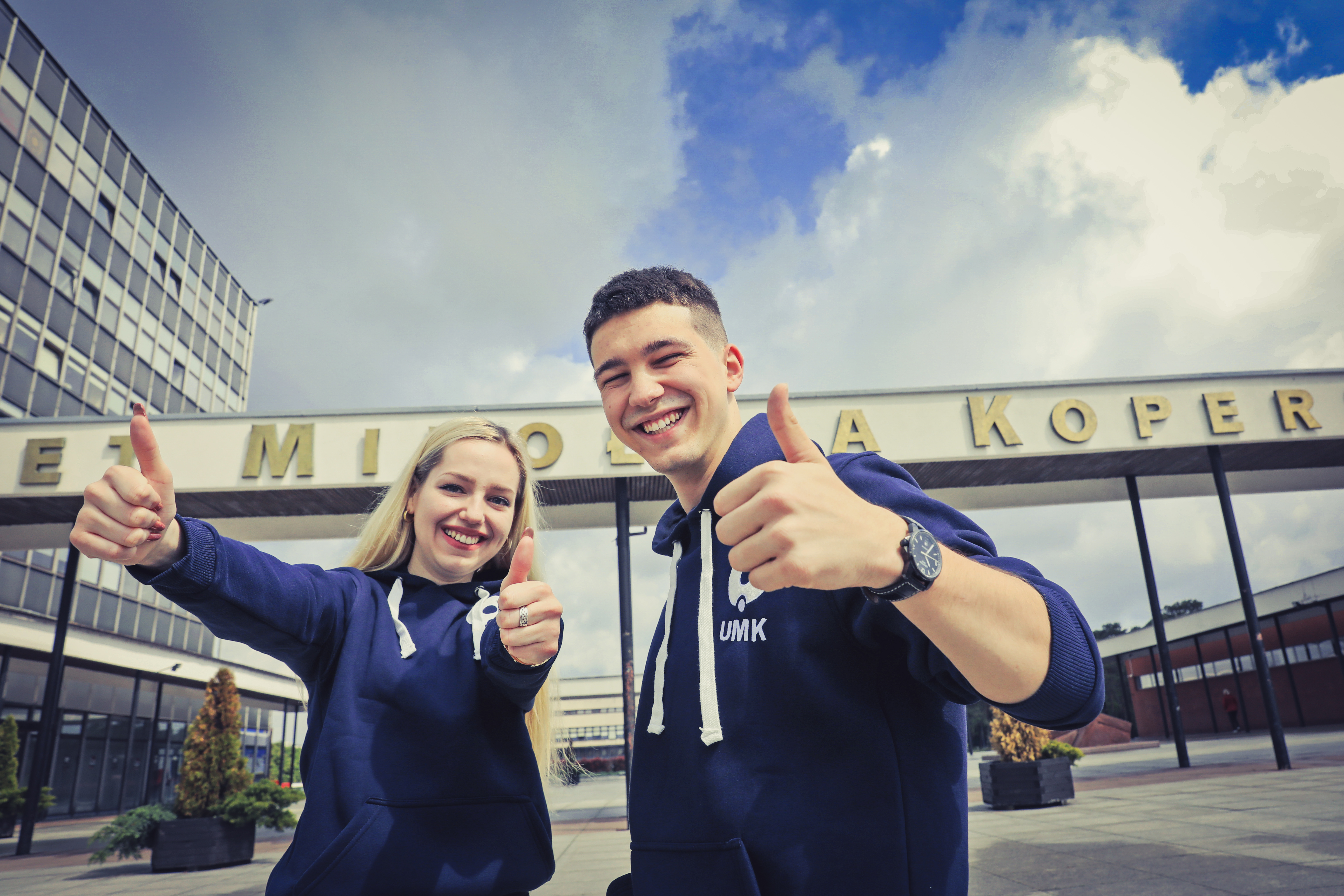 Dwoje studentów przed rektoratem UMK, uśmiechają siei pokazują kciuk w górę