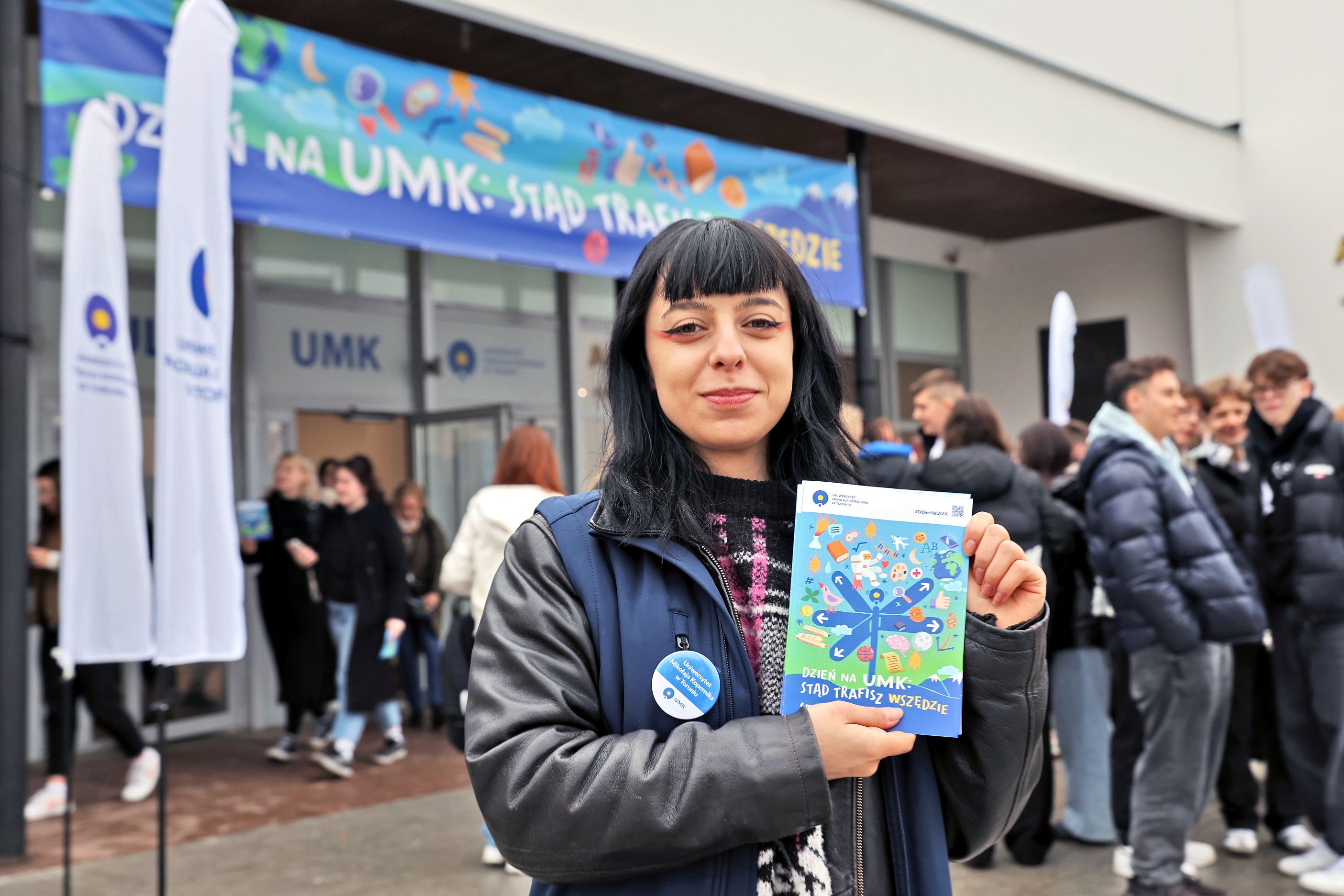 A smiling girl in front of the UMK auditorium holds up a flyer with the slogan &ldquo;UMK: From Here, You Can Go Anywhere&rdquo;