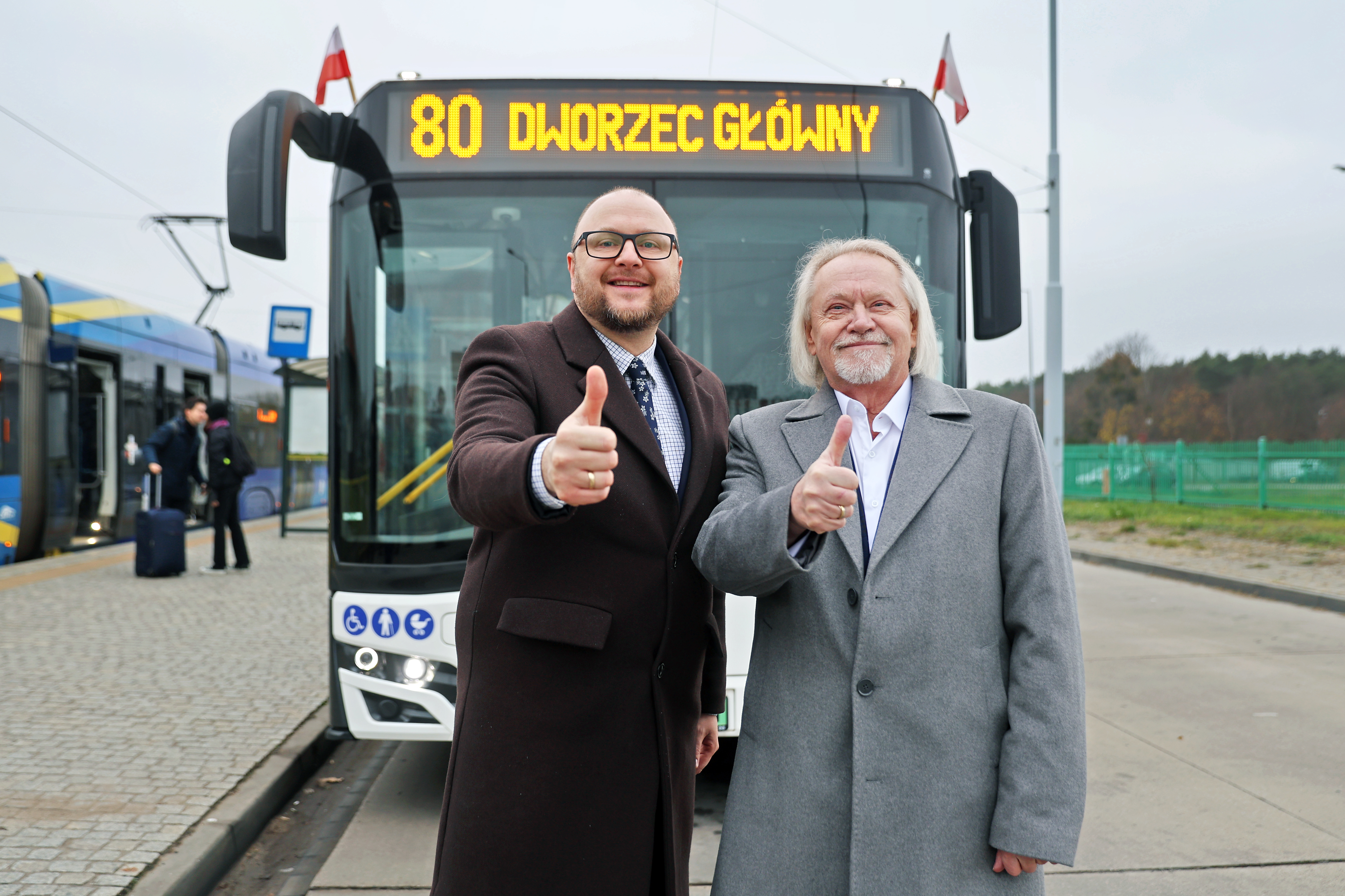 Prezydent Torunia i rektor UMK pokazują kciukw g&oacute;rę, w tle autobus nr 80 z napisem Dworzec Gł&oacute;wny