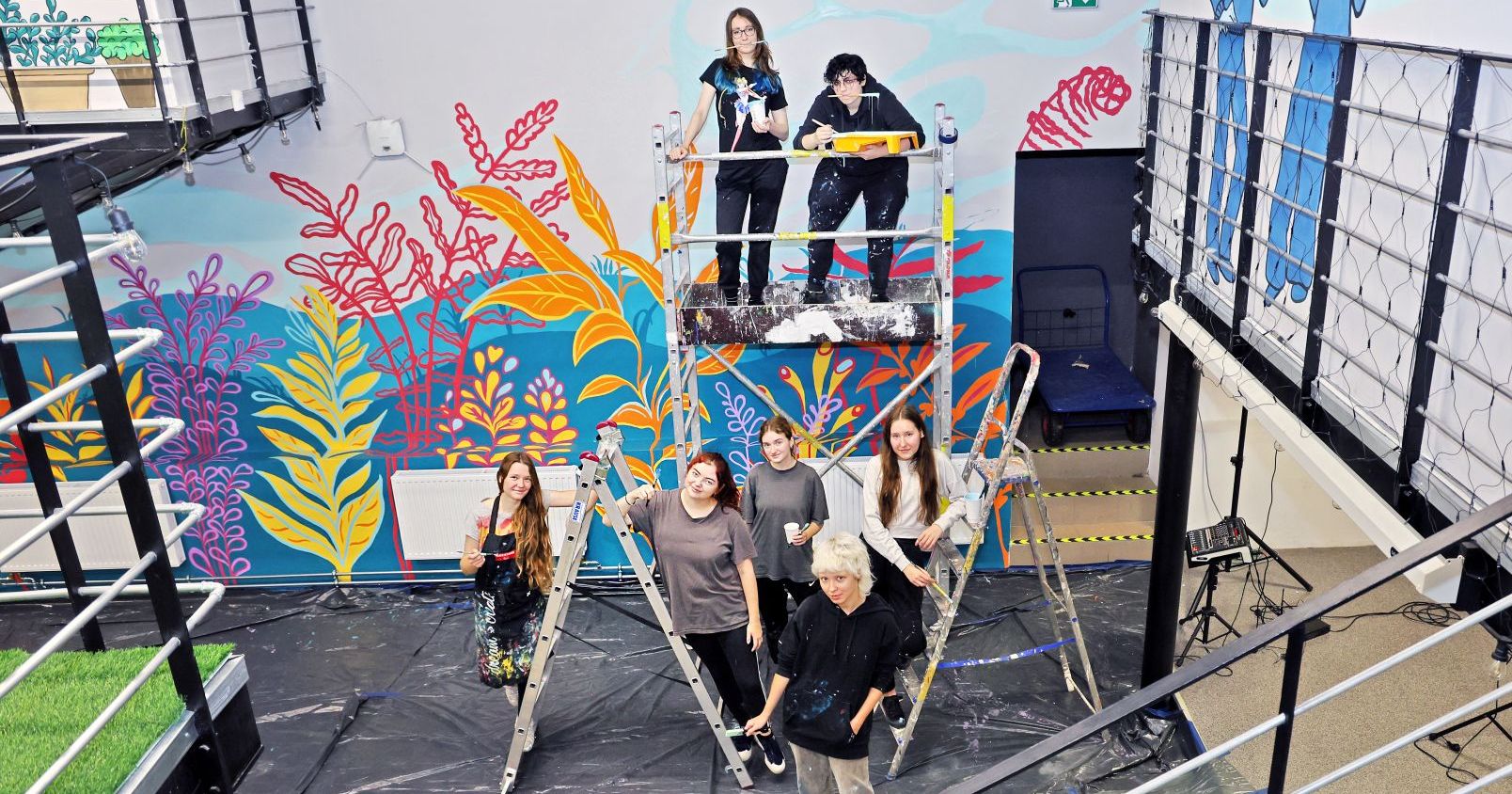 a group of students pose with paintbrushes in front of a colorful mural at Kotłownia