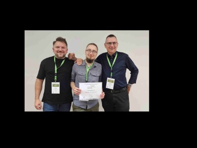 Na zdjęciu znajdują się: dr Szymon Śmiga, mgr Mateusz Witkowski oraz prof. dr hab. Ireneusz Grabowski. Pozują razem na tle jasnej, jednolitej ściany. Wszyscy patrzą w stronę aparatu i uśmiechają się. Mgr Mateusz Witkowski (w środku)  jest ubrany w koszulę w kratę, nosi okulary i ma brodę. Trzyma przed sobą certyfikat. Dwaj pozostali mężczyźni, stojący po jego bokach, obejmują go przyjaźnie ramionami. Wszyscy trzej mają na szyjach zielone smycze z identyfikatorami z konferencji, w której uczestniczą. Dr Szymon Śmiga (po lewej) ma na sobie czarną koszulkę, a prof. dr hab. Ireneusz Grabowski (po prawej) – ciemnoniebieską koszulę i ciemne spodnie. Fotografia ma charakter pamiątkowy i emanuje pozytywną, koleżeńską atmosferą.. Kliknij, aby powiększyć zdjęcie.