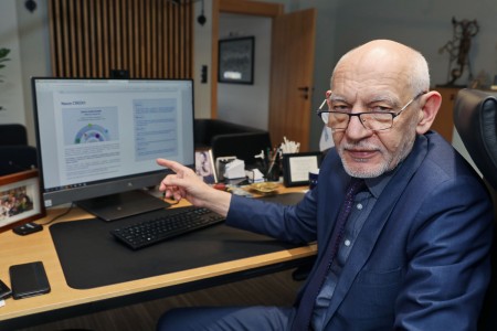 JM Rektor siedzący w gabinecie, przy biurku z komputerem stacjonarnym. Postać odchylona w swoją lewą stronę, w stronę obiektywu, wskazująca palcem ekran komputera z wyświetlonym dokumentem. . Kliknij, aby powiększyć zdjęcie.
