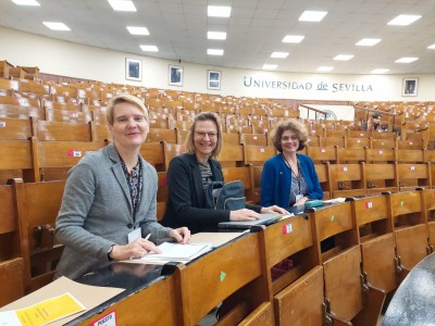 In the audience of a large empty lecture hall, three smiling people are sitting at wooden tables. Click to zoom the picture.