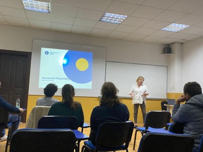 The photo shows the room where classes are held. The teacher explains something to a group of students. In the background there is a slide with a presentation.. Click to zoom the picture.