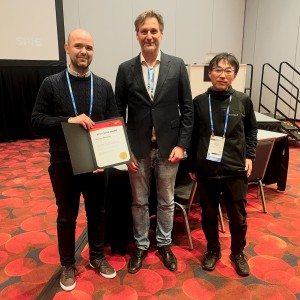 dr inż. Seweryn Morawiec trzyma w ręku dyplom- nagrodę za najlepszą prezentację otrzymaną podczas konferencji, prof. Rainer A. Leitgeb, prof. Yoshiaki Yasuno. Panowie stoją obok siębie na czerwonym dywanie. Ubrani są w małoformalne czarne ubrania. Z tyłu widać stolik z krzesłami.. Kliknij, aby powiększyć zdjęcie.