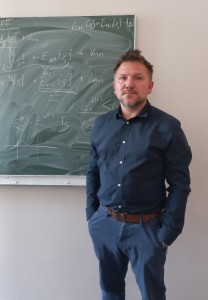 Portret dr. Szymona Śmigi w ciemnoniebieskiej koszuli, stojącego na tle zielonej tablicy zapisanej odręcznymi wzorami naukowymi z zakresu chemii kwantowej.. Kliknij, aby powiększyć zdjęcie.