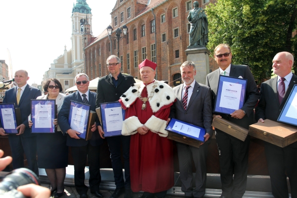 na zdjęciu osiem osób stojących z dyplomami w ramkach, zdjęcie na zewnątrz, przy pomniku Mikołaja Kopernika w Toruniu