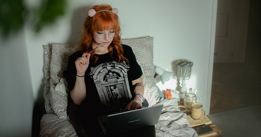 Młoda, rudowłosa kobieta w okularach siedzi na fotelu z europalet, spogląda na ekran laptopa trzymanego na swoich kolanach.