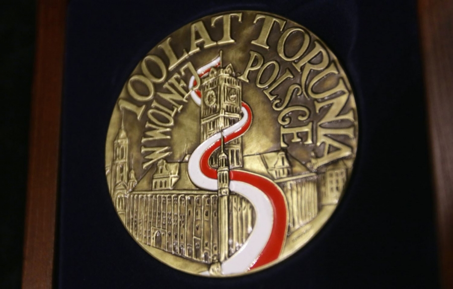 Zdjęcie okrągłego medalu, najpewniej z brązu, z wizerunkem toruńskiego rausza staromiejskiego, z biało-czerwoną flagą spływającą serpentynami z masztu na wieży zegarowej do pomnika Mikołaja Kopernika i napisem: 100 LAT TORUNIA W WOLNEJ POLSCE