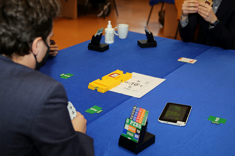 na zdjęciu stół podczas gry w brydża, na którym leżą karty do obstawiania, karty do gry oraz tablet do zapisywania wyników