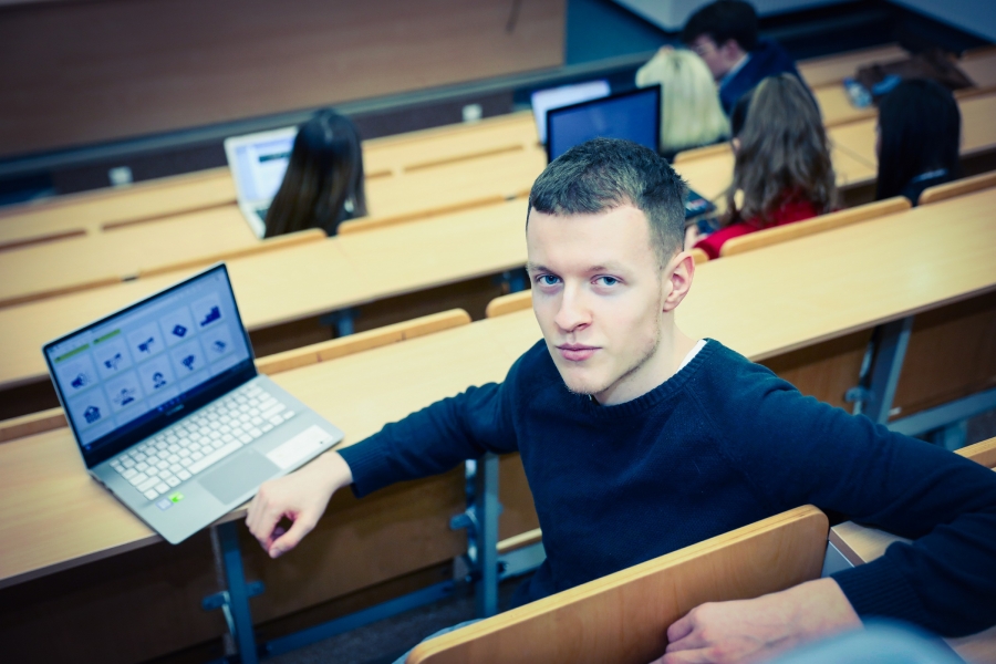 Student patrzący się w obiektyw aparatu. W tle: sala wykładowa oraz laptop, na którym otwarta jest strona rekrutacyjna Uniwersytetu. 