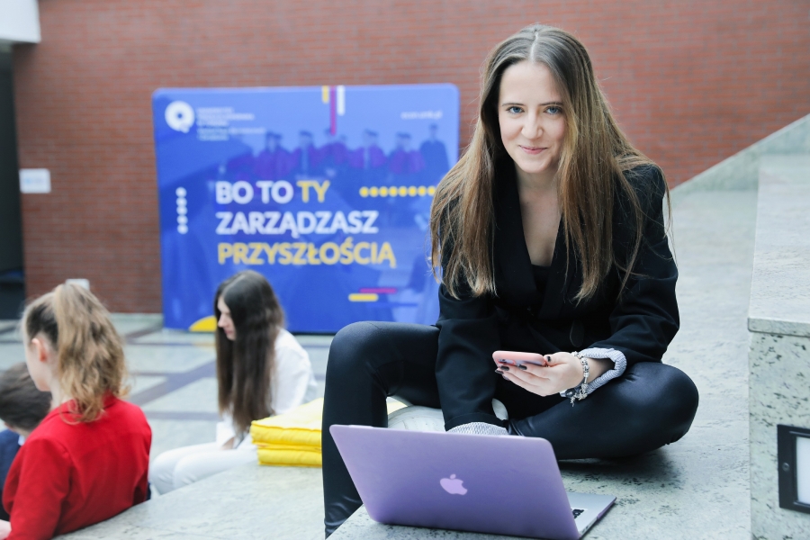 Studentka siedząca na schodach w budynku Wydziału Nauk Ekonomicznych i Zarządzania UMK w Toruniu, patrząca jednocześnie w ekran komputera i telefonu.